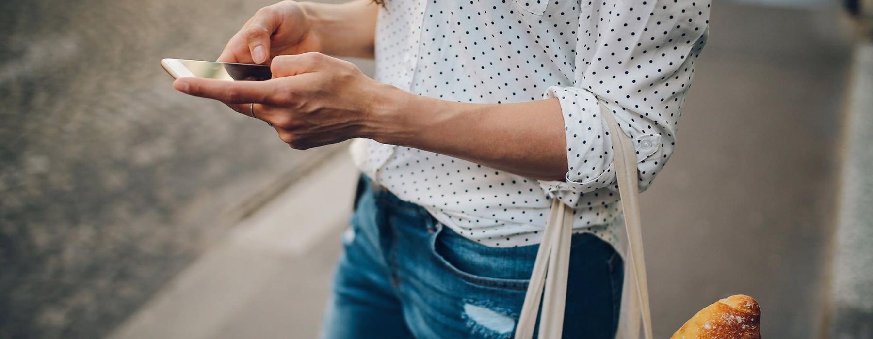 woman walks down the street and texts with a bag of groceries on her arm
